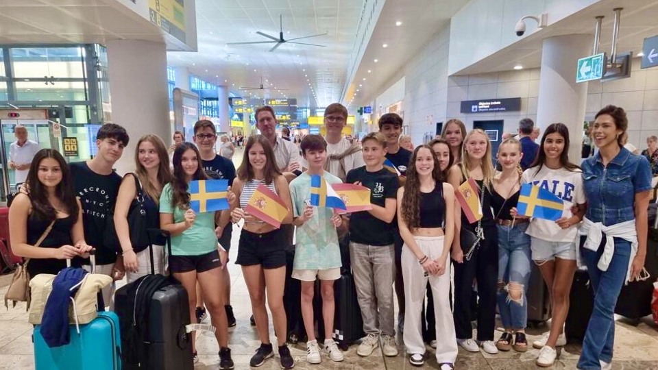 Elever och lärare står på flygplatsen och viftar med svenska och spanska flaggor.